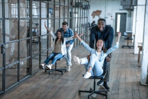 Work hard play hard. Four young cheerful business people in smart casual wear having fun while racing on office chairs and smiling